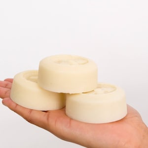 May include: Three round, white, solid soap bars stacked on top of each other in a person's hand.