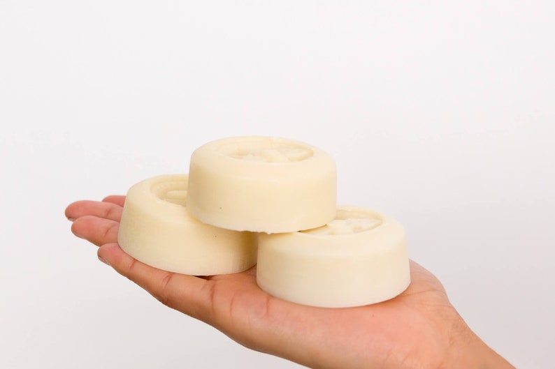 May include: Three round, white, solid soap bars stacked on top of each other in a person's hand.