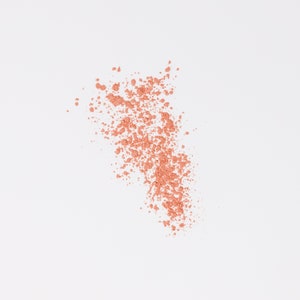 May include: A close-up of a light peach-colored powder scattered on a white background. The powder is in a loose, cloud-like shape.