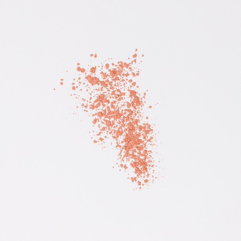 May include: A close-up of a light peach-colored powder scattered on a white background. The powder is in a loose, cloud-like shape.