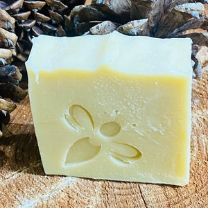 May include: A bar of natural soap with a bee design on the front. The soap is a light yellow color and is sitting on a wooden surface.