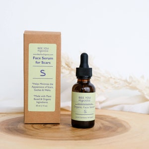 May include: A brown bottle with a dropper filled with a clear liquid. The bottle is labeled "Bee You Organics" and "Organic Face Serum S". The bottle is sitting on a wooden surface.