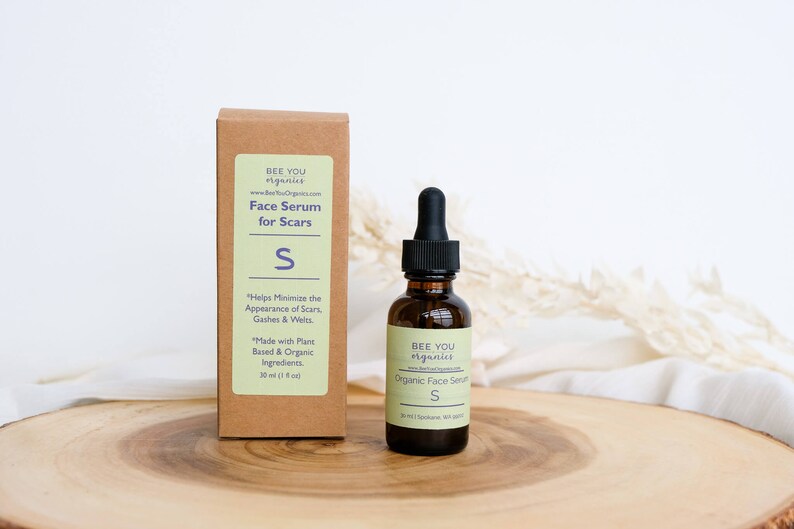 May include: A brown bottle with a dropper filled with a clear liquid. The bottle is labeled "Bee You Organics" and "Organic Face Serum S". The bottle is sitting on a wooden surface.