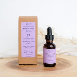 以下が含まれることがあります： 紫色のラベルに「BEE YOU organics Perfect Face Serum P」と製品の利点の説明が書かれた茶色の段ボール箱。箱の隣には、小さな茶色のガラス瓶があり、ドロップボトルには「BEE YOU organics Organic Face Serum P」とラベルが貼られています。