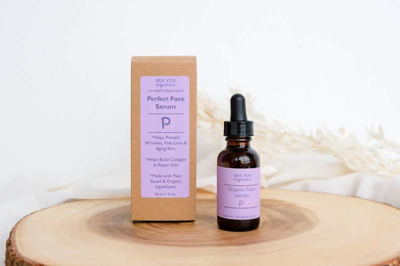 以下が含まれることがあります： 紫色のラベルに「BEE YOU organics Perfect Face Serum P」と製品の利点の説明が書かれた茶色の段ボール箱。箱の隣には、小さな茶色のガラス瓶があり、ドロップボトルには「BEE YOU organics Organic Face Serum P」とラベルが貼られています。