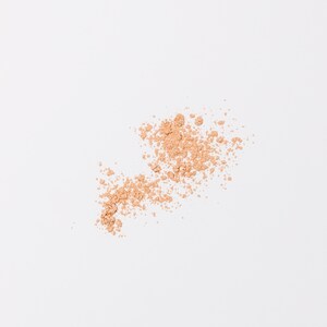 May include: A close-up of a light brown powder scattered on a white surface. The powder is in a loose, irregular shape, suggesting it has been spilled or brushed onto the surface.