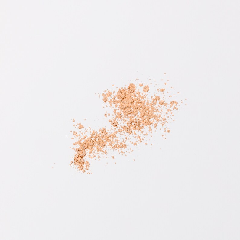 May include: A close-up of a light brown powder scattered on a white surface. The powder is in a loose, irregular shape, suggesting it has been spilled or brushed onto the surface.