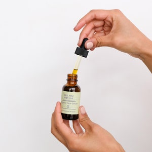 May include: A hand holds a small brown glass bottle with a black dropper. The bottle is labeled "See You Aglow" and "Organic Face Serum".