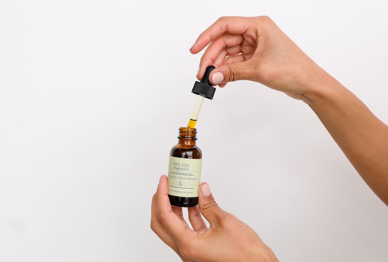 May include: A hand holds a small brown glass bottle with a black dropper. The bottle is labeled "See You Aglow" and "Organic Face Serum".