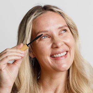 May include: A woman smiles while applying mascara to her eyelashes. She is holding a black mascara wand with a wooden handle.