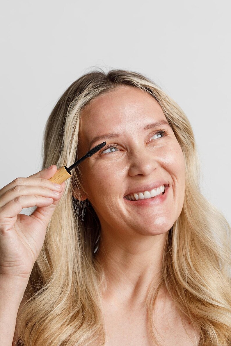 May include: A woman smiles while applying mascara to her eyelashes. She is holding a black mascara wand with a wooden handle.