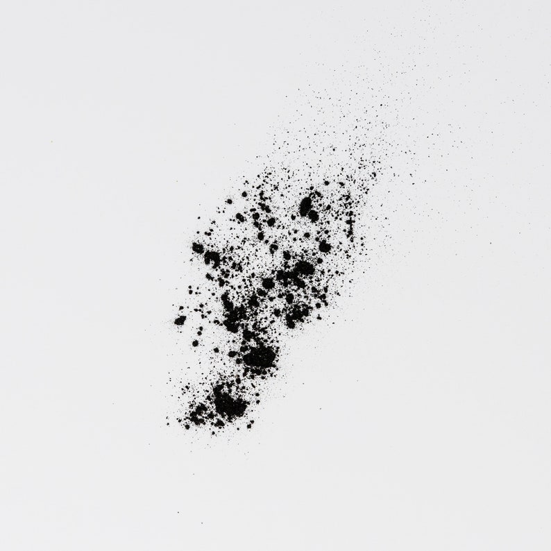 May include: Black powder scattered on a white background. The powder is in a loose, irregular shape, with some of it forming small clumps.