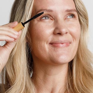 May include: A woman applies mascara to her eyelashes with a black mascara wand. The mascara wand is attached to a wooden tube.