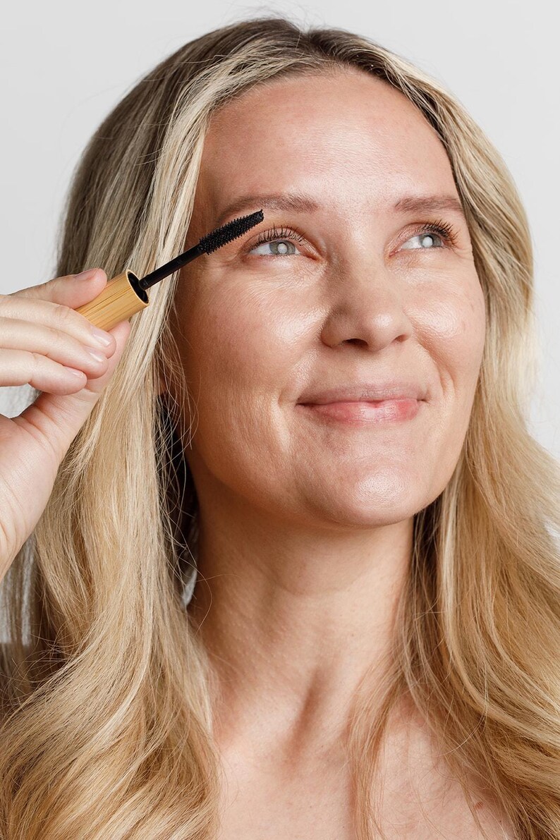 May include: A woman applies mascara to her eyelashes with a black mascara wand. The mascara wand is attached to a wooden tube.