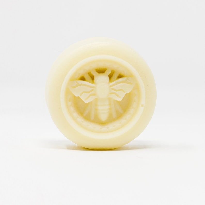 May include: A round, white soap bar with a bee design embossed on the front.