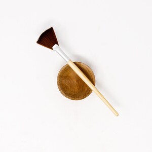 May include: A wooden bowl with a brown makeup brush with a fan-shaped brush head.