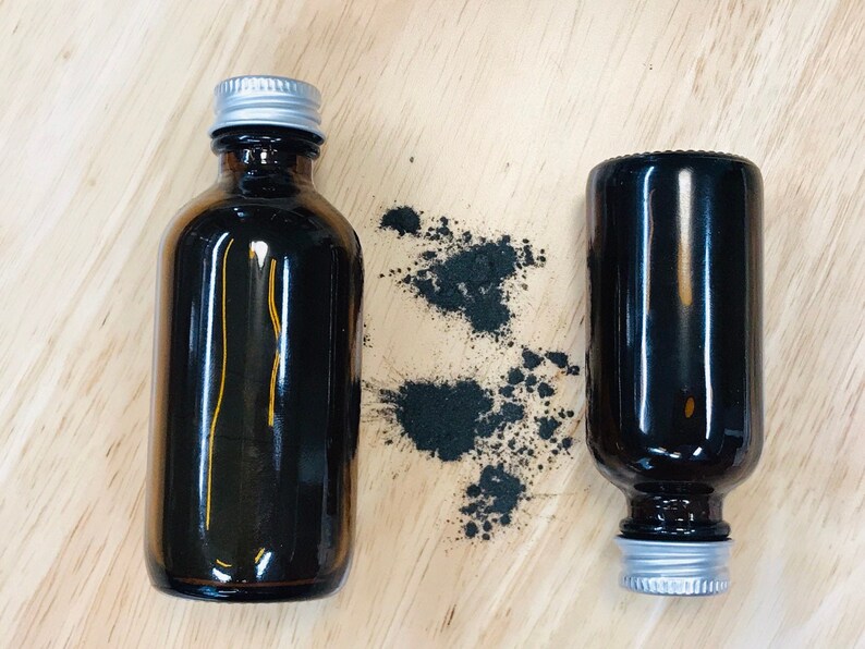 May include: Two amber glass bottles with silver lids, one upright and one lying down, on a wooden surface. Black powder is scattered around the upright bottle.