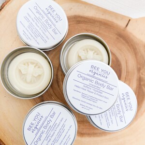 May include: Four silver tins containing organic body bars with a bee logo. The tins are labeled "Bee You Organics" and "Organic Body Bar" with different scents: Lavender, Unscented, and Dream.
