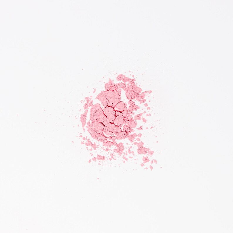 May include: A close-up of a pink powder blush makeup product on a white background.