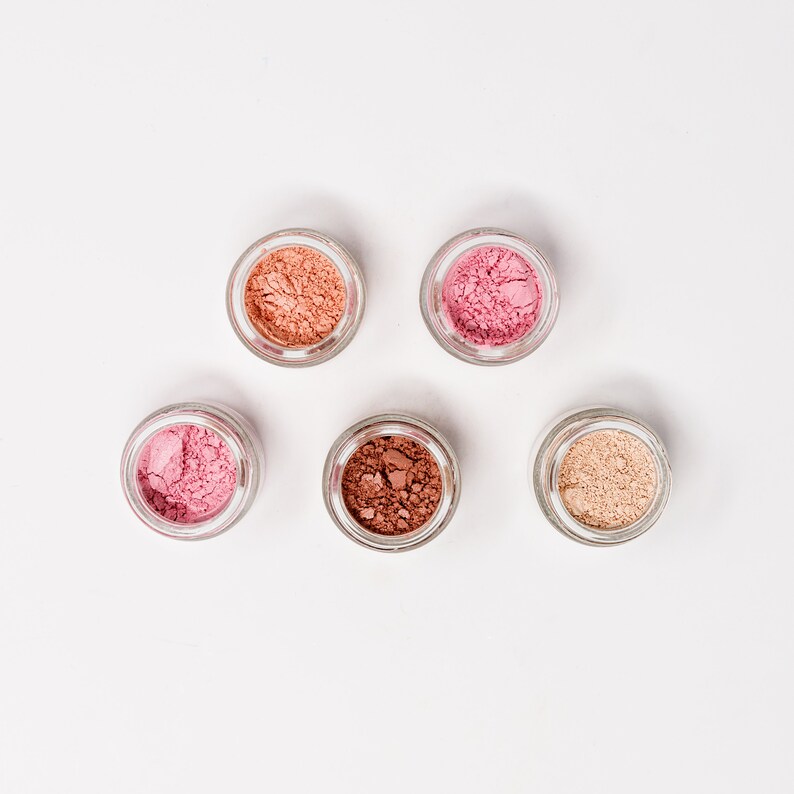 May include: Five small glass jars with lids containing different shades of pink, brown, and beige powder. The jars are arranged in a circle on a white background.