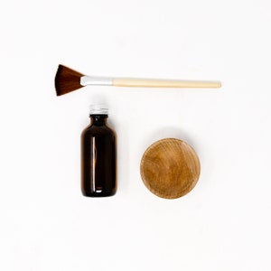 May include: A brown glass bottle with a silver lid, a wooden bowl, and a fan-shaped brush with a wooden handle on a white background.