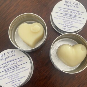 May include: Two silver tins with lids containing heart-shaped organic body bars. The tins are labeled "Bee You Organics" and "Organic Body Bar" with the scent "Orange Vanilla Dream". The text on the tin reads "Softens, Hydrates, Helps Heal, Repair & Protect Skin. Gently massage into clean, dry skin. Spokane, WA 99202 net wt .25 oz"