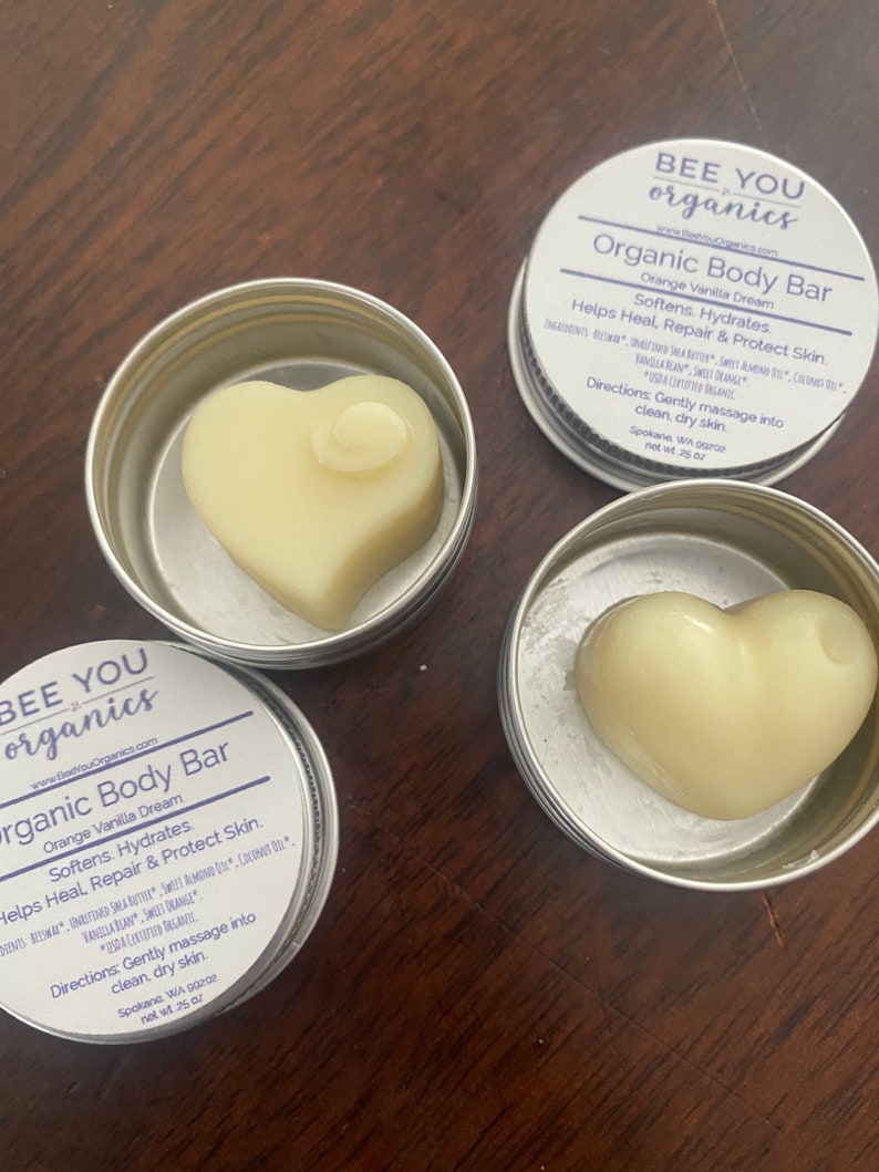May include: Two silver tins with lids containing heart-shaped organic body bars. The tins are labeled "Bee You Organics" and "Organic Body Bar" with the scent "Orange Vanilla Dream". The text on the tin reads "Softens, Hydrates, Helps Heal, Repair & Protect Skin. Gently massage into clean, dry skin. Spokane, WA 99202 net wt .25 oz"