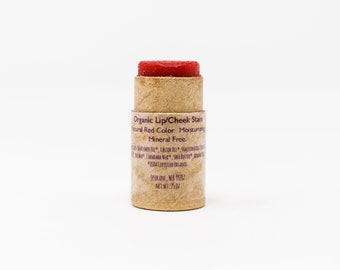 May include: A brown cardboard tube with a red lip and cheek stain. The tube has a label that reads "Organic Lip/Cheek Stain Natural Red Color. Moisturizing Mineral Free. USDA Certified Organic Spokane, WA 91202 Net Wt: .75 oz"