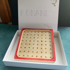 May include: A wooden game board with a red border and a pattern of small circles. The board is in a white box with the word "KONANE" written on the lid. The box also has a small dolphin logo.