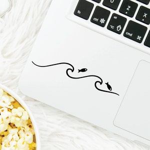 Op de afbeelding: Een witte laptop met een zwart toetsenbord en een zwart-witte sticker met een golf en een vis op de bovenkant van de laptop. De laptop staat op een wit, pluche oppervlak.