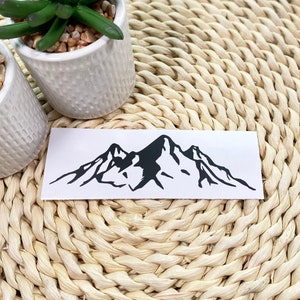 May include: A black and white mountain range silhouette sticker on a white background. The sticker is on a woven straw surface.
