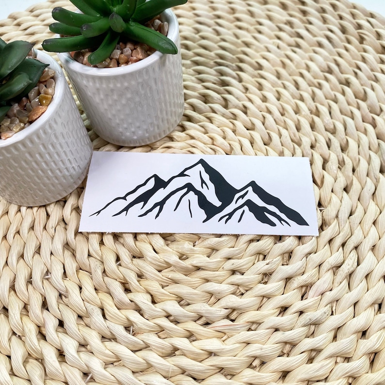 May include: A black and white mountain range silhouette sticker on a white background. The sticker is on a woven straw mat.