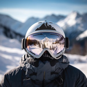 May include: A white ski helmet with a black mountain range sticker on the top. The helmet has a pair of goggles with a reflective lens that shows a mountain range reflected in the lens.