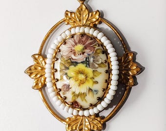 Peut inclure: Un pendentif ovale présentant une peinture florale centrale avec des fleurs jaunes et roses sur fond crème. Le centre est entouré d'une bordure de petites perles blanches et enchâssé dans un cadre en métal doré avec des motifs de feuilles.