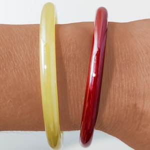 May include: Two smooth, circular bangle bracelets worn together. One bangle is a translucent, pale yellow color, while the other is a deep, glossy red. Both bracelets feature a simple, rounded profile with a polished finish and no additional embellishments.