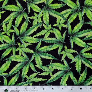 May include: A fabric featuring a repeating pattern of green cannabis leaves on a black background, positioned above a white Johnson measuring tape that shows a length of 10 inches, providing a clear scale for the printed botanical design.