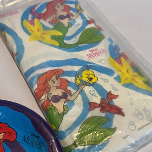 May include: A white plastic tablecloth with a blue and white swirl pattern. The tablecloth features images of Ariel, Flounder, Sebastian, and a starfish from Disney's The Little Mermaid. The text "Disney's The Little Mermaid" is printed on the tablecloth.