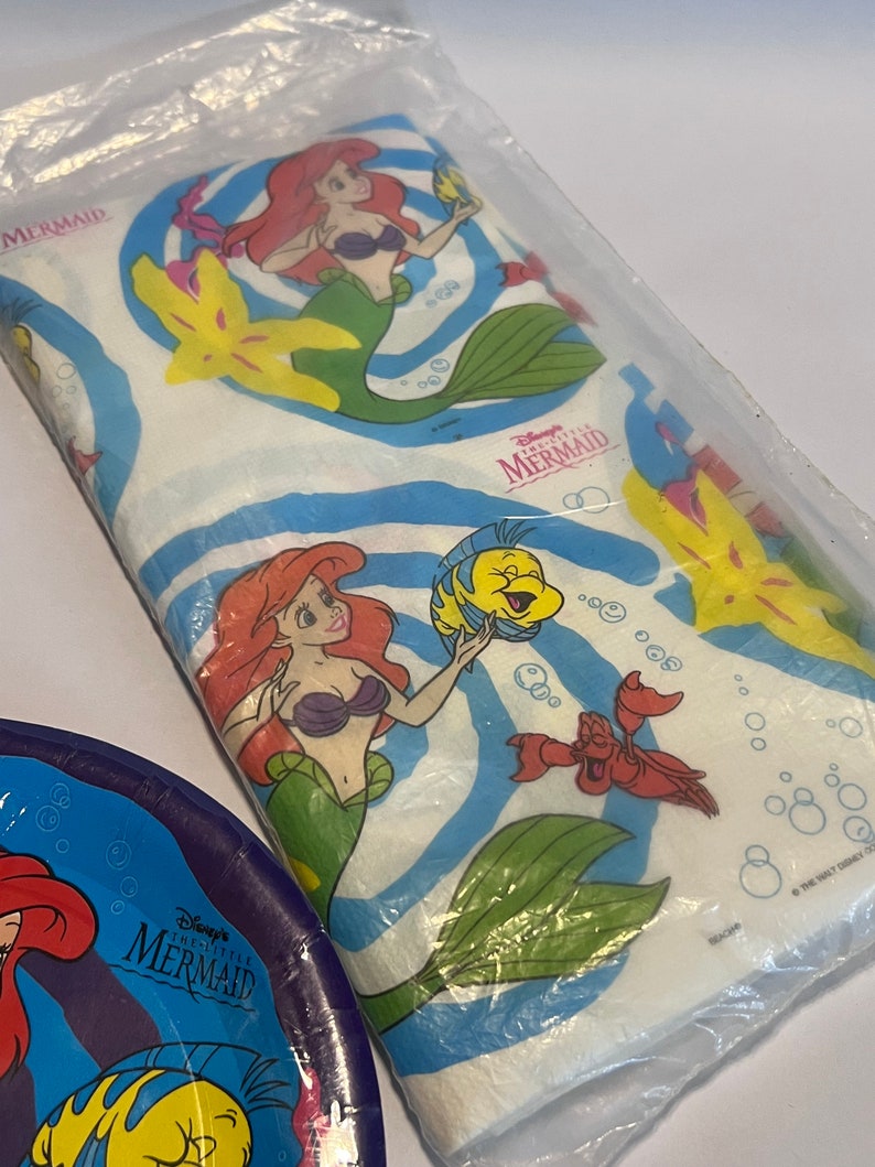 May include: A white plastic tablecloth with a blue and white swirl pattern. The tablecloth features images of Ariel, Flounder, Sebastian, and a starfish from Disney's The Little Mermaid. The text "Disney's The Little Mermaid" is printed on the tablecloth.