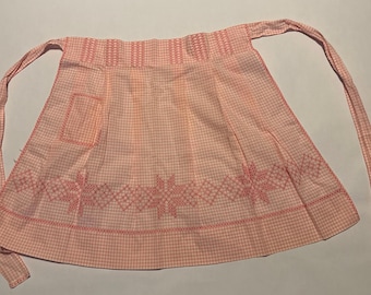 Op de afbeelding: Een vintage roze en witte gingham schort met een zak en decoratieve borduursels. De schort heeft lange strikken en een gedetailleerd geborduurd ontwerp langs de onderrand. De schort is gemaakt van een lichte stof.