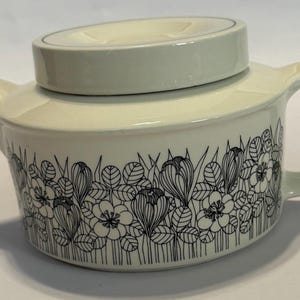 May include: Cream-colored ceramic teapot with a black floral pattern around the base. The teapot has a rounded body, a curved handle, and a lid with a small, raised section. The spout is short and angled.