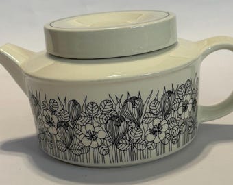 May include: Cream-colored ceramic teapot with a black floral pattern around the base. The teapot has a rounded body, a curved handle, and a lid with a small, raised section. The spout is short and angled.