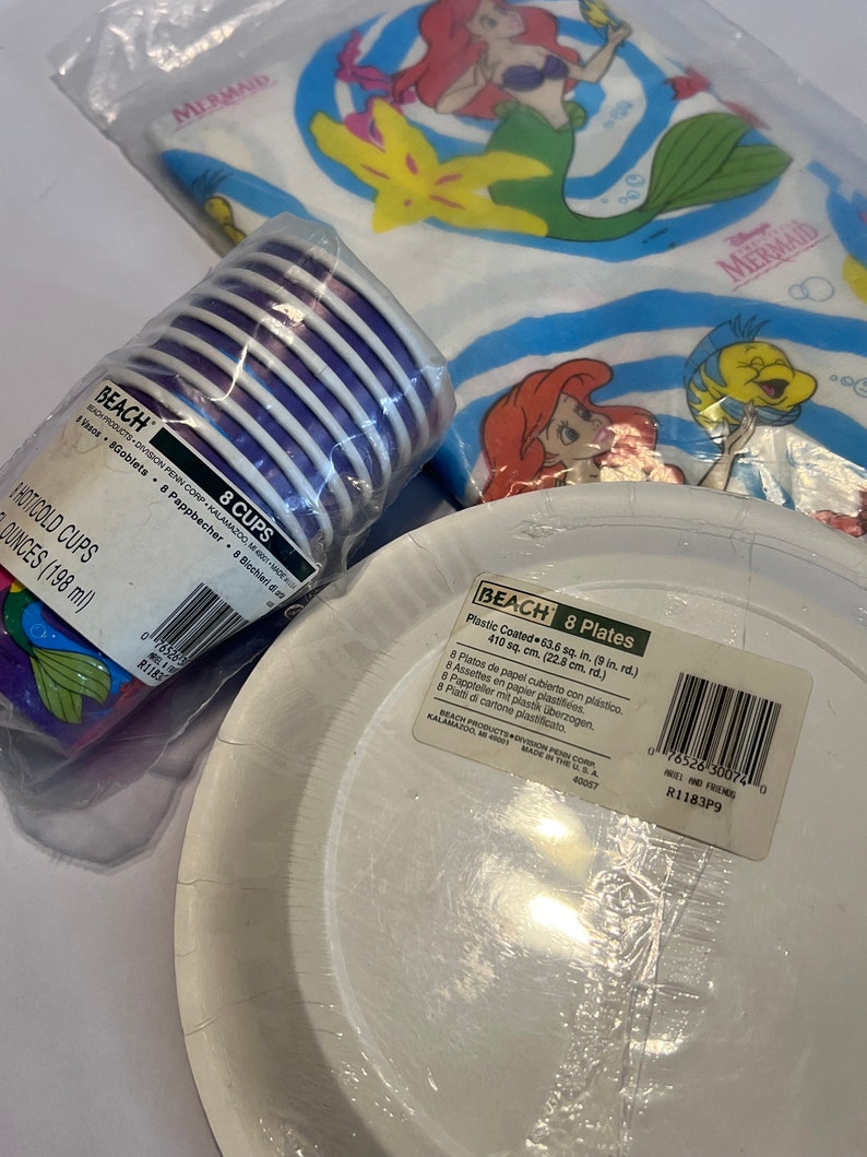 May include: A set of 8 purple and white paper cups and 8 white paper plates with a plastic coating. The plates are 22.8 cm in diameter. The packaging features a Disney Little Mermaid theme with images of Ariel and Flounder.