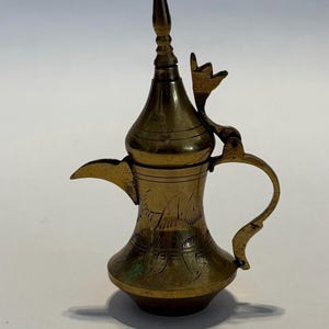 May include: A small, ornate brass coffee pot with a long spout, curved handle, and a decorative finial on the lid. The pot features etched designs and a unique, stylized handle. The overall design suggests a Middle Eastern or antique aesthetic.