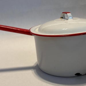Może przedstawiać: Biały emaliowany rondel z czerwonym uchwytem i obramowaniem. Rondel ma pokrywkę z czerwonym akcentem na uchwycie. Garnek jest przeznaczony do gotowania i ma klasyczny, vintage wygląd.