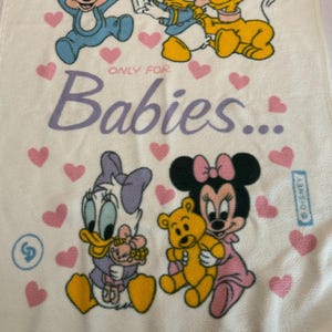 Puede incluir: Manta de bebé blanca con los personajes de Disney Mickey Mouse, Donald Duck, Pluto, Daisy Duck y Minnie Mouse. La manta tiene corazones rosas y el texto "ONLY FOR Babies..." en morado.