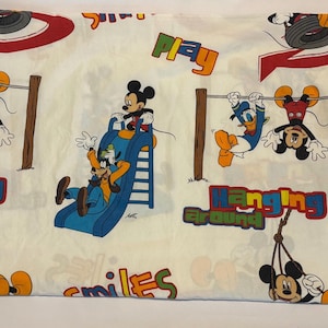Puede incluir: S&aacute;bana de cama blanquecina con los personajes de dibujos animados Mickey Mouse, Donald Duck y Goofy. La s&aacute;bana tiene un dise&ntilde;o l&uacute;dico con las palabras "Play", "Hanging Around" y "Smiles" en letras coloridas. Los personajes est&aacute;n representados en un parque infantil.