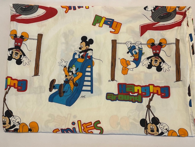 Puede incluir: S&aacute;bana de cama blanquecina con los personajes de dibujos animados Mickey Mouse, Donald Duck y Goofy. La s&aacute;bana tiene un dise&ntilde;o l&uacute;dico con las palabras "Play", "Hanging Around" y "Smiles" en letras coloridas. Los personajes est&aacute;n representados en un parque infantil.