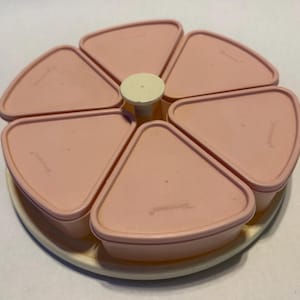 Puede incluir: Una bandeja de servicio Tupperware rosa vintage con seis compartimentos triangulares y tapas. La bandeja tiene una base blanca y un asa central blanca. Los compartimentos están dispuestos en un patrón circular.