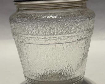 May include: A clear glass jar with a white lid. The jar is shaped like a barrel and has a textured surface.