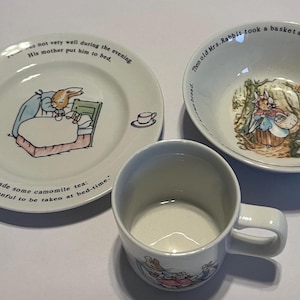 May include: A set of three Peter Rabbit themed children's dishes. The set includes a small bowl, a plate, and a mug. The bowl and plate feature illustrations of Peter Rabbit and the text "The old Mrs. Rabbit took a basket and her umbrella. And went through the wood until she came to the other side." The plate also features the text "He was not very well during the evening. His mother put him to bed. And made some camomile tea: One table-spoonful to be taken at bed-time."